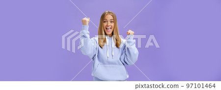 Cheerful happy and supportive young girlfriend with fair hair and tan in warm hoodide raising fists in cheer and triumph smiling saying yeah celebrating goal or success over purple background 97101464