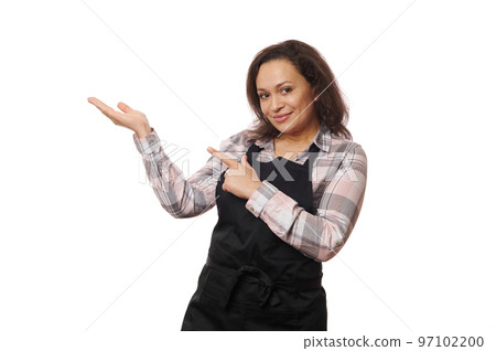Pleasant African American woman waitress, barmaid wearing black chef's apron, smiling on camera and pointing at copy advertising space she's holding on her hand palm up, isolated on a white background 97102200