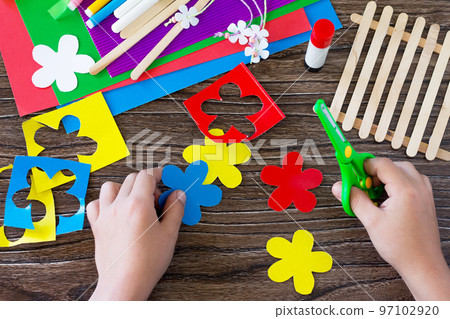 A child is cutting details of a gift of summer flowers on a wooden sticks fence. Handmade. Project of children's creativity, handicrafts, crafts for kids. 97102920
