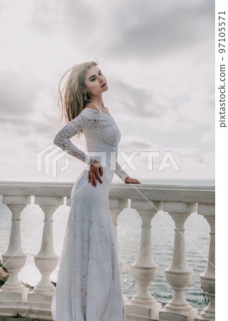 Romantic bride, blond girl in white wedding dress with open shoulders posing on open terrace with backdrop of the sea and rocks. Stylish young woman standing on terrace and looking on ocean at sunset 97105571