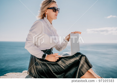 Digital nomad, Business woman working on laptop by the sea. Pretty lady typing on computer by the sea at sunset, makes a business transaction online from a distance. Freelance, remote work on vacation 97105583