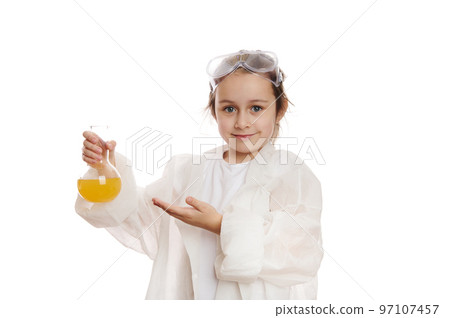 Adorable little child girl, smart schoolgirl, young chemist-scientist at chemistry class, points at laboratory flask with liquid chemical, looking at camera isolated on white background. Copy ad space 97107457