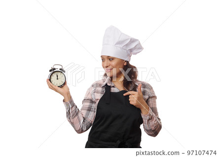 Happy waitress, pastry chef, baker confectioner or new catering establishment owner, in black apron points at black alarm clock showing the opening and closing time of cafeteria, on white background 97107474