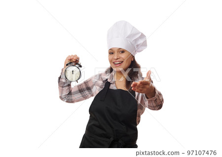 Charming female waitress in a chef's hat and black apron, holding an alarm clock in her hands and smiling, extends her hand to the camera, inviting to visit a new open catering establishment Charming female waitress in a chef's hat and black apron, holding an alarm clock in her hands and smiling, extends her hand to the camera, inviting to visit a new open catering establishment 97107476