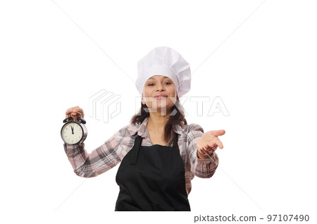 Female pastry chef in black apron, shows alarm clock with the opening time of a coffee shop or bakery, extends her hand, invite you to visit a new catering establishment, isolated on white background Female pastry chef in black apron, shows alarm clock with the opening time of a coffee shop or bakery, extends her hand, invite you to visit a new catering establishment, isolated on white background 97107490