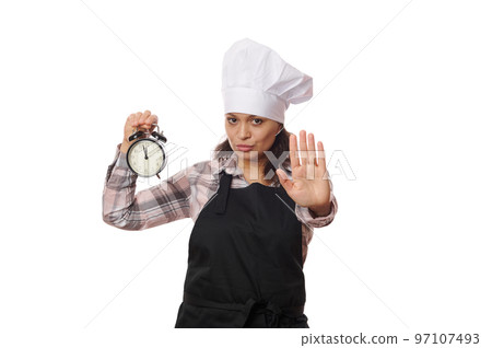 Serious multi-ethnic woman waitress, cafe worker in black apron holding black alarm clock showing opening and closing time, extending her hand to show stop or prohibition gesture, on white background. 97107493