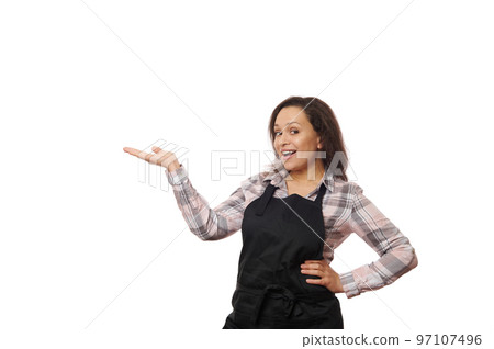 Cheerful smiling multiethnic brunette woman, female bartender, barmaid, waitress, in black apron, selling hot drink coffee, tea, holding a copy space for your advertising, isolated on white background 97107496