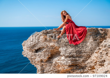 A girl with flowing hair in a long red dress sits on a rock above the sea. The stone can be seen in the sea. A girl with flowing hair in a long red dress sits on a rock above the sea. The stone can be seen in the sea. 97109024