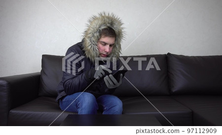 A young man is sitting at home in a winter jacket and trying to keep warm. A man sits without heating and calls on the phone and asks for help. 97112909