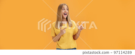 Amused enthusiastic and charismatic girl with blond hair and tanned pure skin without makeup pointing and looking at upper left corner with joyful happy smile over orange background 97114289