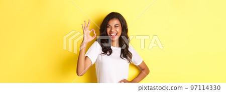 Portrait of confident and attractive african-american girl, have everything under control, showing okay sign and winking to assure you, approve something good, posing over yellow background Portrait of confident and attractive african-american girl, have everything under control, showing okay sign and winking to assure you, approve something good, posing over yellow background 97114330