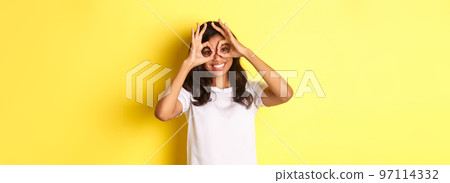 Image of happy and funny african-american girl, making finger glasses and looking through them, standing over yellow background Image of happy and funny african-american girl, making finger glasses and looking through them, standing over yellow background 97114332