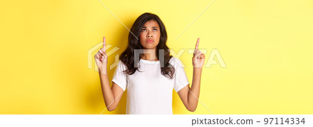 Portrait of disappointed and sad african-american girl, pointing and looking up, frowning displeased, standing gloomy over yellow background 97114334