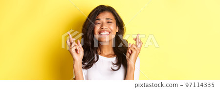 Close-up of nervous african-american girl, crossing fingers for good luck and waiting for important results, making a wish, standing over yellow background Close-up of nervous african-american girl, crossing fingers for good luck and waiting for important results, making a wish, standing over yellow background 97114335