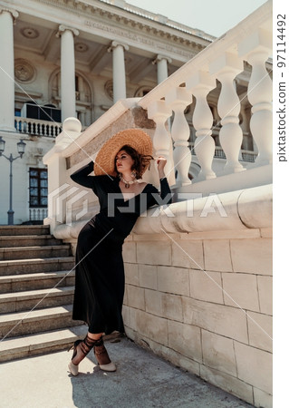 Fashion photo of a beautiful model in an elegant hat and black dress posing against the backdrop of a building with columns. 97114492