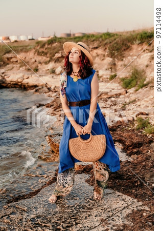 A woman in a dress, hat and with a straw bag is standing on the beach enjoying the sea. Happy summer holidays 97114498