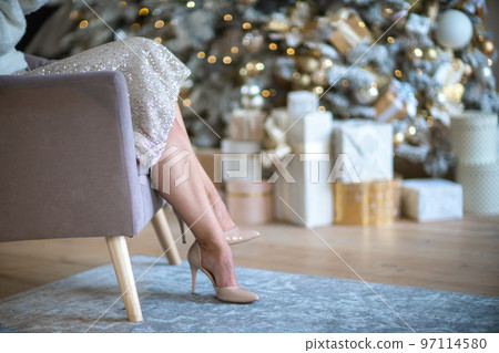 Women's legs. Cropped close-up photo of healthy beautiful elegant female legs in beige high-heeled shoes, woman sitting in a chair on the background of a Christmas tree with gifts 97114580