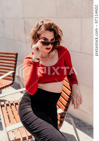 Portrait of a woman on the street. An attractive woman in glasses, a red blouse and a black skirt is sitting on a bench outside. Portrait of a woman on the street. An attractive woman in glasses, a red blouse and a black skirt is sitting on a bench outside. 97114602