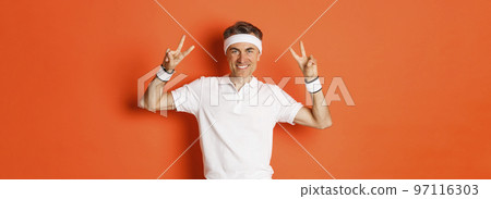 Portrait of handsome, healthy middle-aged male athlete in sportswear, showing peace signs and smiling, workout in gym, standing over orange background Portrait of handsome, healthy middle-aged male athlete in sportswear, showing peace signs and smiling, workout in gym, standing over orange background 97116303