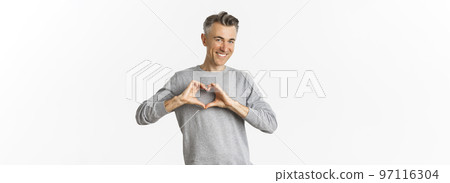 Portrait of cheeky middle-aged man in grey sweater, smiling and showing heart sign, love someone, standing over white background Portrait of cheeky middle-aged man in grey sweater, smiling and showing heart sign, love someone, standing over white background 97116304