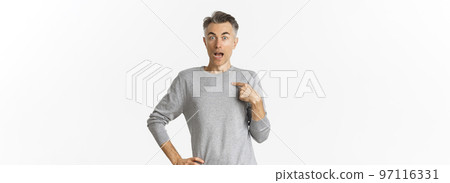 Portrait of handsome middle-aged gay man looking surprised, pointing at himself, being chosen, standing over white background in grey basic sweater 97116331
