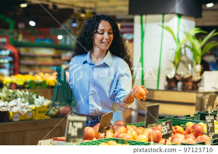 Smiling beautiful Latin American business woman chooses and buys fresh fruits in supermarket for restaurant, home. He is holding apples and an eco bag. 97123710