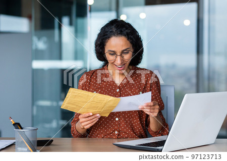 Happy young Latin American woman received bill with earned salary, cash reward, good news. He is sitting at the desk in the office, holding an envelope with a letter in his hands. Happy young Latin American woman received bill with earned salary, cash reward, good news. He is sitting at the desk in the office, holding an envelope with a letter in his hands. 97123713