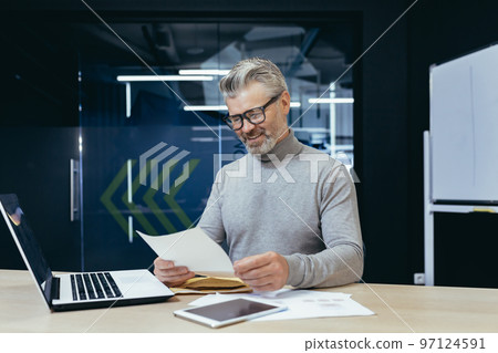Mature successful gray haired businessman smiling and happy received letter notification, senior male boss working inside office using laptop reading good news in letter. Mature successful gray haired businessman smiling and happy received letter notification, senior male boss working inside office using laptop reading good news in letter. 97124591