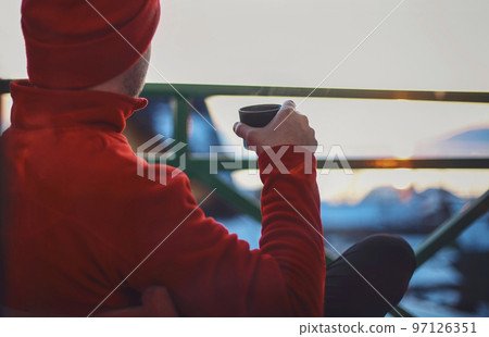 Man in winter clothing drinking hot herbal tea. Winter evening from a beautiful balcony in the suset time on Baikal Lake. 97126351