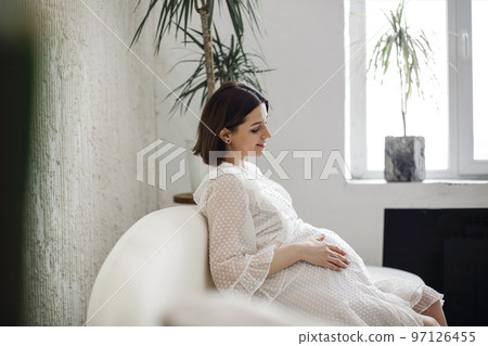 Beautiful charming stylish pregnant woman with dark hair sitting on a white sofa in a beige vintage dress Beautiful charming stylish pregnant woman with dark hair sitting on a white sofa in a beige vintage dress 97126455
