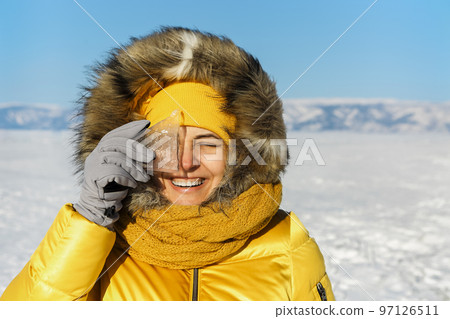 Happy woman wearing winter yellow jacket holding piece of transparent crushed ice cubes over her face 97126511