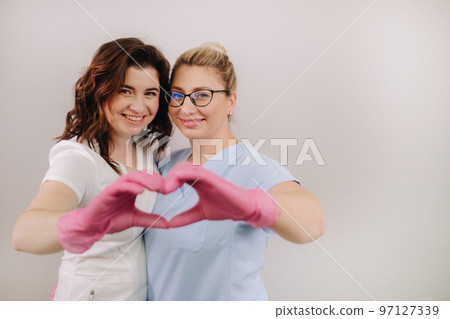 Portrait of two qualified cosmetologists-women, confidently smiling and making a heart with their hands in a medical center Portrait of two qualified cosmetologists-women, confidently smiling and making a heart with their hands in a medical center 97127339