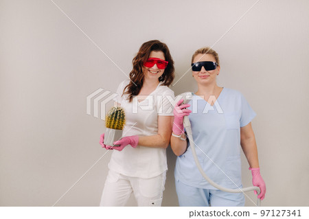 Two cosmetologists in protective glasses are holding a laser depilation device and a cactus in their hands Two cosmetologists in protective glasses are holding a laser depilation device and a cactus in their hands 97127341