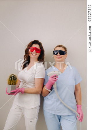Two cosmetologists in protective glasses are holding a laser depilation device and a cactus in their hands Two cosmetologists in protective glasses are holding a laser depilation device and a cactus in their hands 97127342