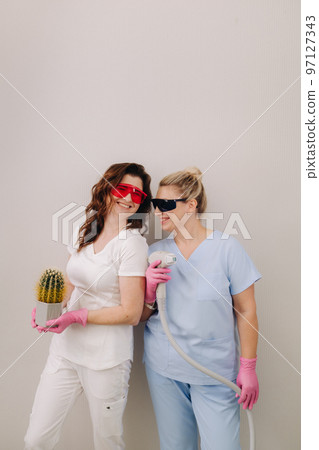 Two cosmetologists in protective glasses are holding a laser depilation device and a cactus in their hands 97127343