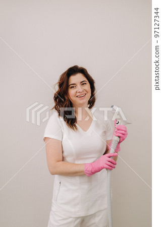 Portrait of the master of laser hair removal. A woman in a white coat holds a laser hair removal device in her hands Portrait of the master of laser hair removal. A woman in a white coat holds a laser hair removal device in her hands 97127344