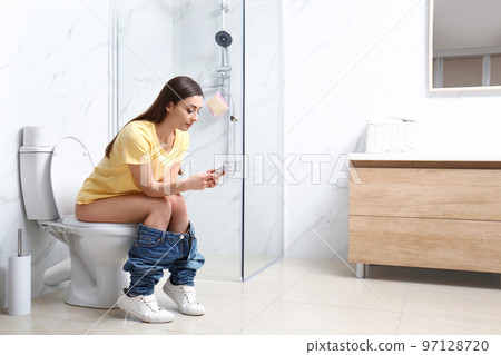 Woman with smartphone sitting on toilet bowl in bathroom Woman with smartphone sitting on toilet bowl in bathroom 97128720