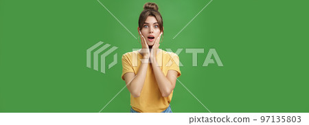 Woman feeling surprised and delighted enjoying good result after detox and applying facial skincare mask touching cheeks opening mouth from amazement and excited posing over green background 97135803