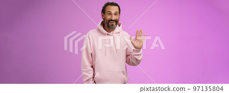 Cheeky charismatic funny happy smiling mature man bearded grey hair in pink hoodie waving palm hello nice meet you greetng gesture welcoming guests say hi standing purple background friendly relaxed 97135804