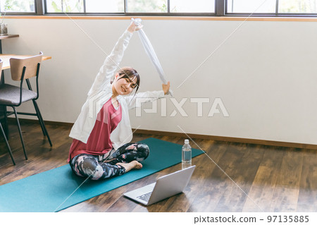 Asian woman doing light exercise, stretching, yoga, online lessons using a towel and a computer in the living room at home Asian woman doing light exercise, stretching, yoga, online lessons using a towel and a computer in the living room at home 97135885