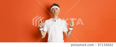 Portrait of handsome middle-aged male athlete, wearing sports clothing, looking and pointing fingers up at promo banner, standing against orange background Portrait of handsome middle-aged male athlete, wearing sports clothing, looking and pointing fingers up at promo banner, standing against orange background 97138882