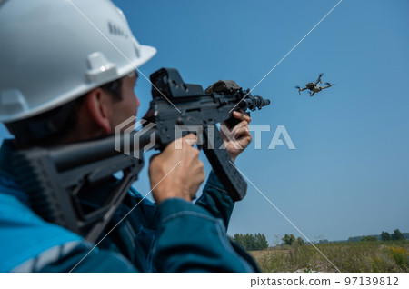 Caucasian man in a helmet shoots a flying drone with a rifle. Caucasian man in a helmet shoots a flying drone with a rifle. 97139812
