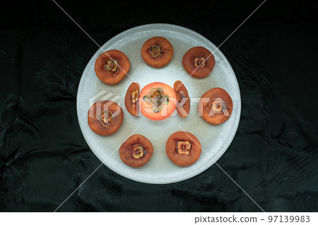 One fresh ripe persimmon fruit with Dried persimmons and colorful leaves on round ceramic tray. One fresh ripe persimmon fruit with Dried persimmons and colorful leaves on round ceramic tray. 97139983