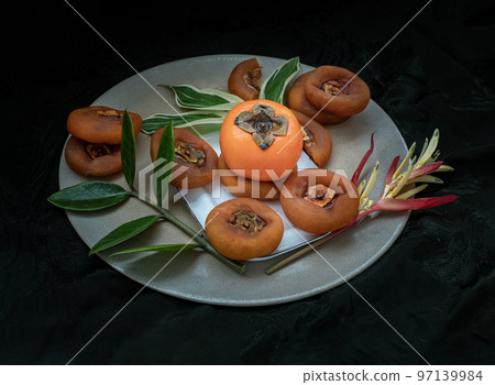 One fresh ripe persimmon fruit with Dried persimmons and colorful leaves on round ceramic tray. 97139984