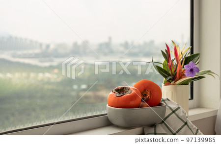 Few fresh ripe persimmons fruits in the ceramic bowl beside with Colorful flower vase. Few fresh ripe persimmons fruits in the ceramic bowl beside with Colorful flower vase. 97139985
