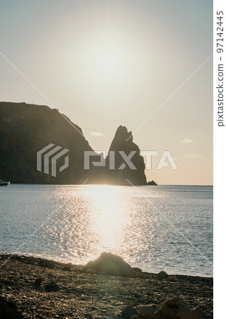A red burning sunset over the sea with rocky volcanic cliff. Abstract nature summer ocean sea background. Small waves on golden warm water surface with bokeh lights from sun. A red burning sunset over the sea with rocky volcanic cliff. Abstract nature summer ocean sea background. Small waves on golden warm water surface with bokeh lights from sun. 97142445