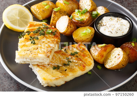 Portion of grilled cod fish with baked jacket potatoes, creamy sauce and lemon close-up in a plate. Horizontal Portion of grilled cod fish with baked jacket potatoes, creamy sauce and lemon close-up in a plate. Horizontal 97150260