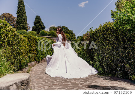 a beautiful woman with long brown hair and long white dress runs along a path along beautiful bushes in the park rear view 97152109