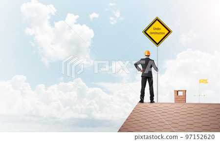 Young businessman on house brick roof holding yellow signboard. Mixed media Young businessman on house brick roof holding yellow signboard. Mixed media 97152620