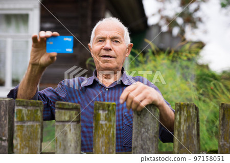 Portrait of satisfied old man 70s with gray hair smiling while holding credit card over country house background 97158051